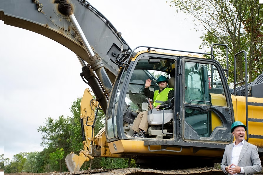 Matt Cooney sits in an excavator at a groundbreaking event for Walton Oaks. Oakland County Executive Dave Coulter is at bottom right.